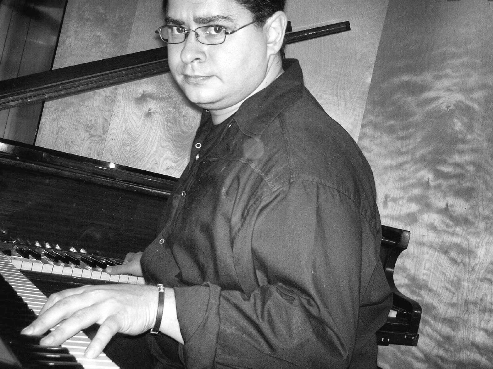 Yan Kayary con camisa negra y lentes, con las manos sobre un piano acústico