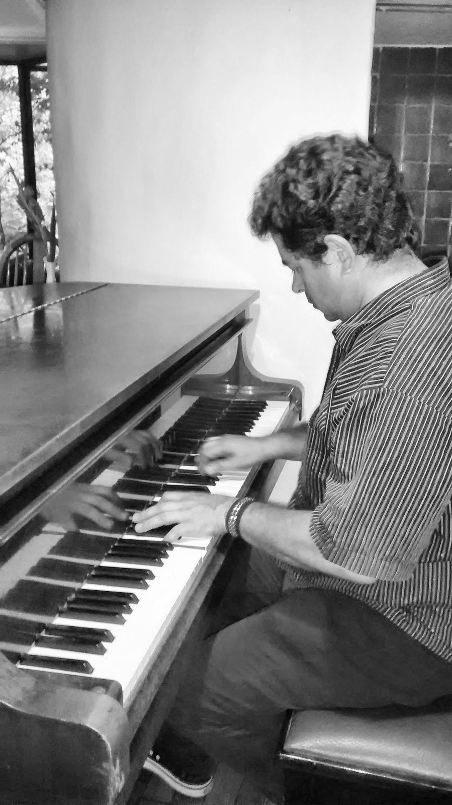 Yan Kayary con camisa azul, tocando un piano vertical de madera junto a una ventana