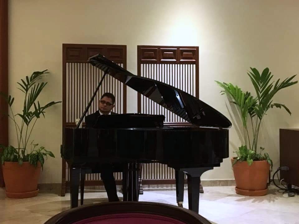 Piano de cola negro entre plantas y un biombo, con el pianista al fondo